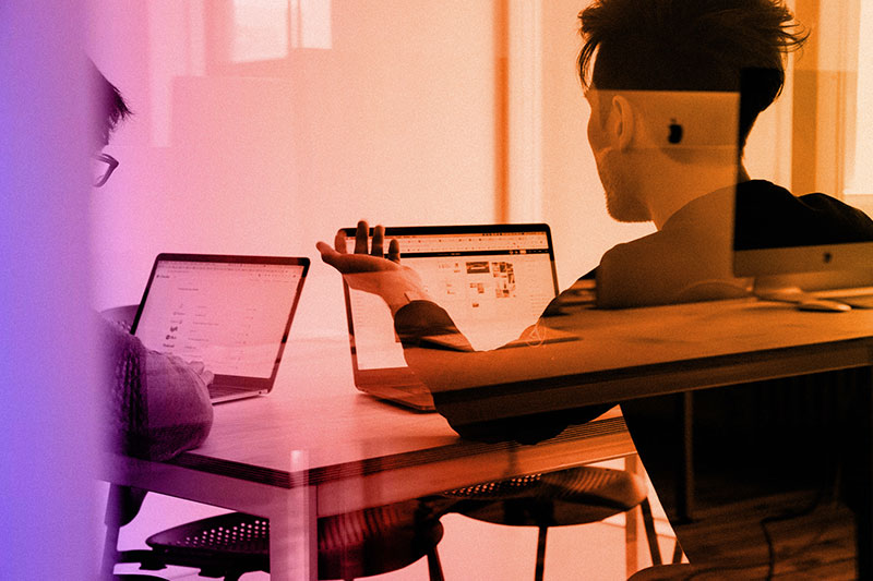 Two people sit at a table with open laptops, engaged in discussion. One person gestures toward their screen, possibly explaining or presenting something. The image is taken through glass, adding reflections of the room and computers.