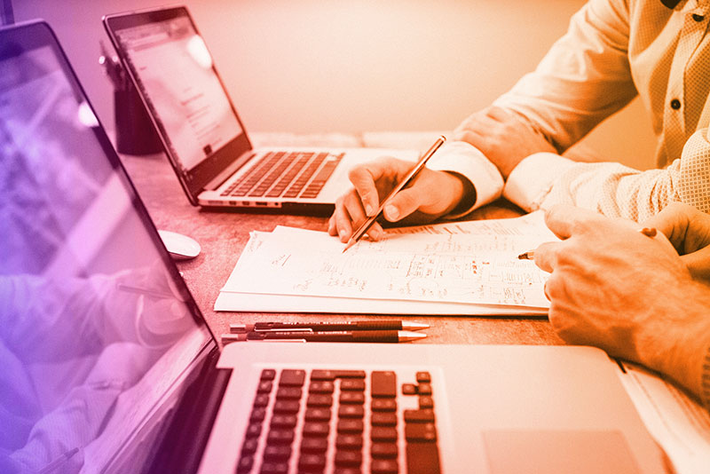 Two people collaborating at a desk with open laptops, reviewing and writing on printed documents. One person is holding a pen and pointing at the paper, suggesting discussion or planning. The workspace is modern and focused on productivity.